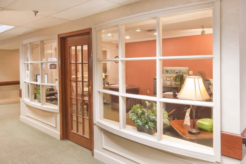 View of an office area inside a senior living facility seen through large glass windows with white frames. The office has a wooden door with glass panels, a desk with a lamp, a green plant, and various office items. The walls inside the office are painted a warm reddish color, and the hallway outside has beige walls and carpeted flooring.