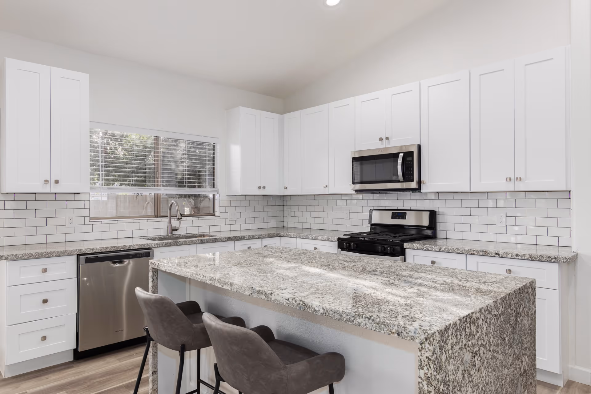 Modern kitchen with white cabinets, granite countertops, a large kitchen island with two gray bar stools, stainless steel dishwasher, stove, and microwave. A window with blinds is above the sink.