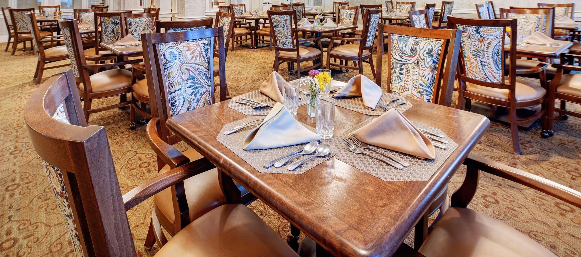 A dining room with multiple wooden tables and chairs. Each table is set with beige cloth napkins, silverware, glasses, and small flower vases. The chairs have patterned fabric backs and leather seats. The room has a carpeted floor with a decorative pattern.