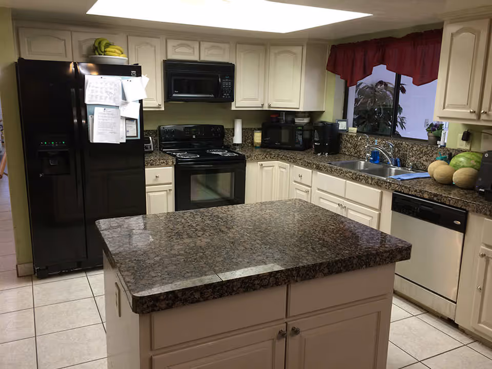 A kitchen with cream-colored cabinets and granite countertops. The kitchen features a black refrigerator with papers attached, a black stove with an overhead microwave, a double sink under a window with a red valance, a dishwasher, a microwave, a coffee maker, and a kitchen island with a granite countertop in the center.