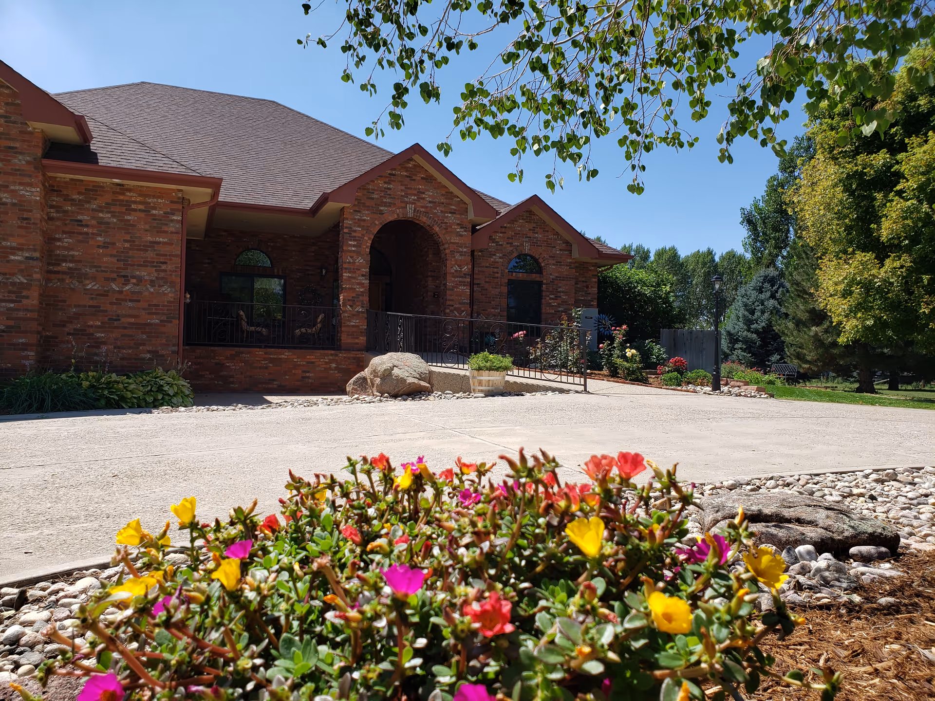 Front exterior view of a brick building with a sloped roof, arched entryway, and a ramp leading to the entrance. In the foreground, colorful flowers and landscaping rocks are visible under a clear blue sky.