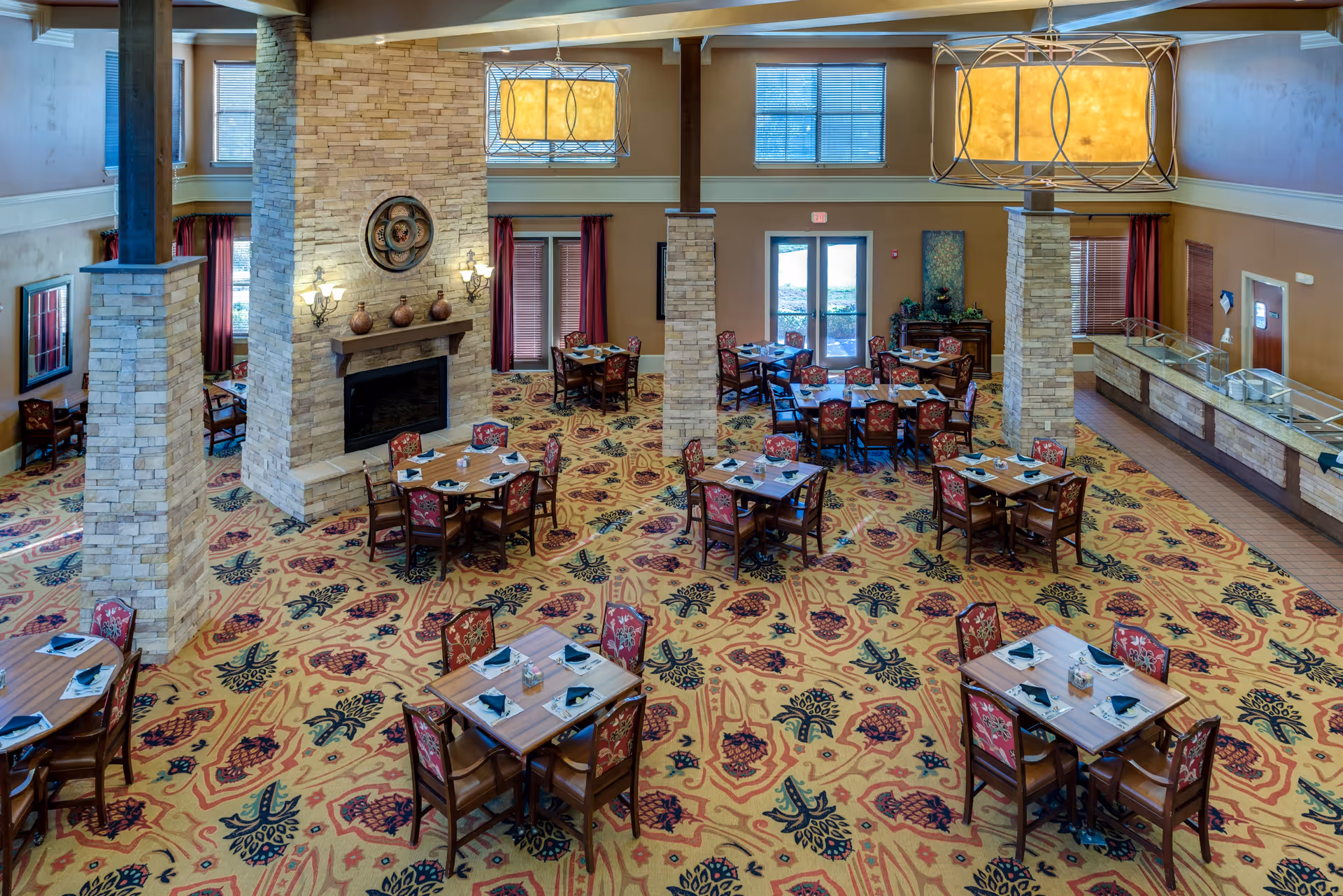 Spacious dining room with multiple wooden tables and chairs arranged neatly on a patterned carpet. The room features a large stone fireplace in the center, decorative wall sconces, large windows with blinds, and two large hanging light fixtures. There is a serving area with a counter on the right side of the room.