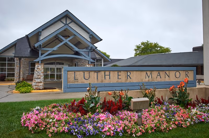 Front entrance of Luther Manor with a stone-and-timber porte-cochere, a large sign reading "LUTHER MANOR", and colorful flowerbeds in the foreground.