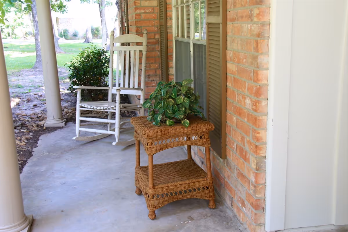 A porch area with a white wooden rocking chair, a wicker side table with a green potted plant on top, and a brick wall with a window and brown shutters. The porch has concrete flooring and is supported by white columns. There is greenery and trees visible in the background.