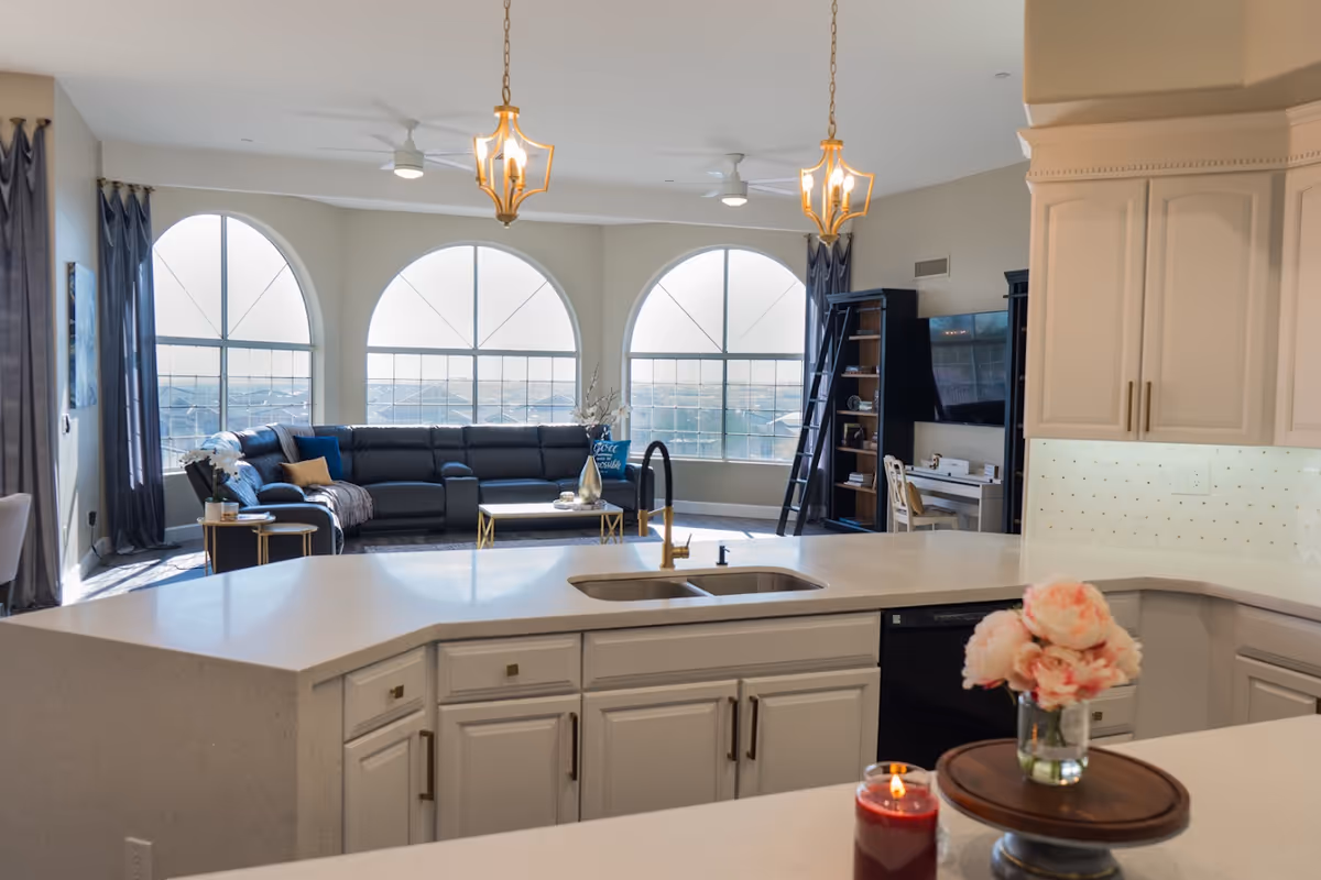 Bright and spacious open-concept living area with a modern kitchen in the foreground featuring a large white countertop with a sink, a vase with pink flowers, and a lit red candle. In the background, there is a cozy living room with a large dark sectional sofa, a coffee table, tall arched windows with gray curtains, and a black entertainment center with a TV and bookshelf.
