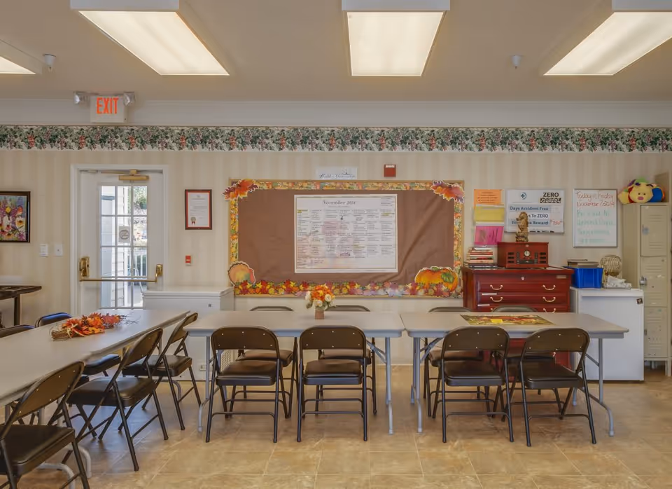 A senior living facility common room with folding tables and chairs arranged in rows. The walls are decorated with a floral border and a large bulletin board displaying a calendar and various notices. There is a door with an exit sign above it, a small refrigerator, lockers, and a wooden cabinet with books and decorations on top. The room is well-lit with ceiling lights.