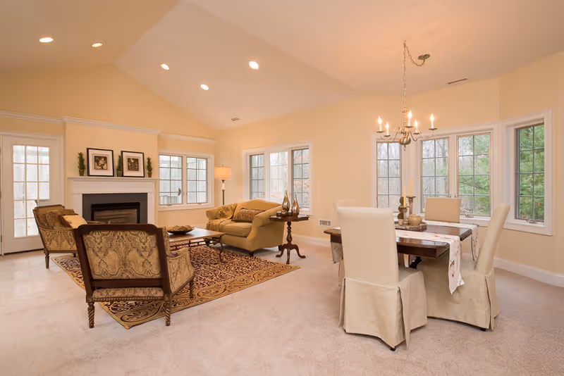 A bright and spacious living and dining area with beige walls and carpet. The living area features a fireplace with framed artwork above it, two upholstered armchairs, a sofa, a wooden coffee table on a patterned rug, and a floor lamp. The dining area has a wooden table with a white table runner and four chairs with beige slipcovers, illuminated by a chandelier. Large windows provide natural light and views of trees outside.