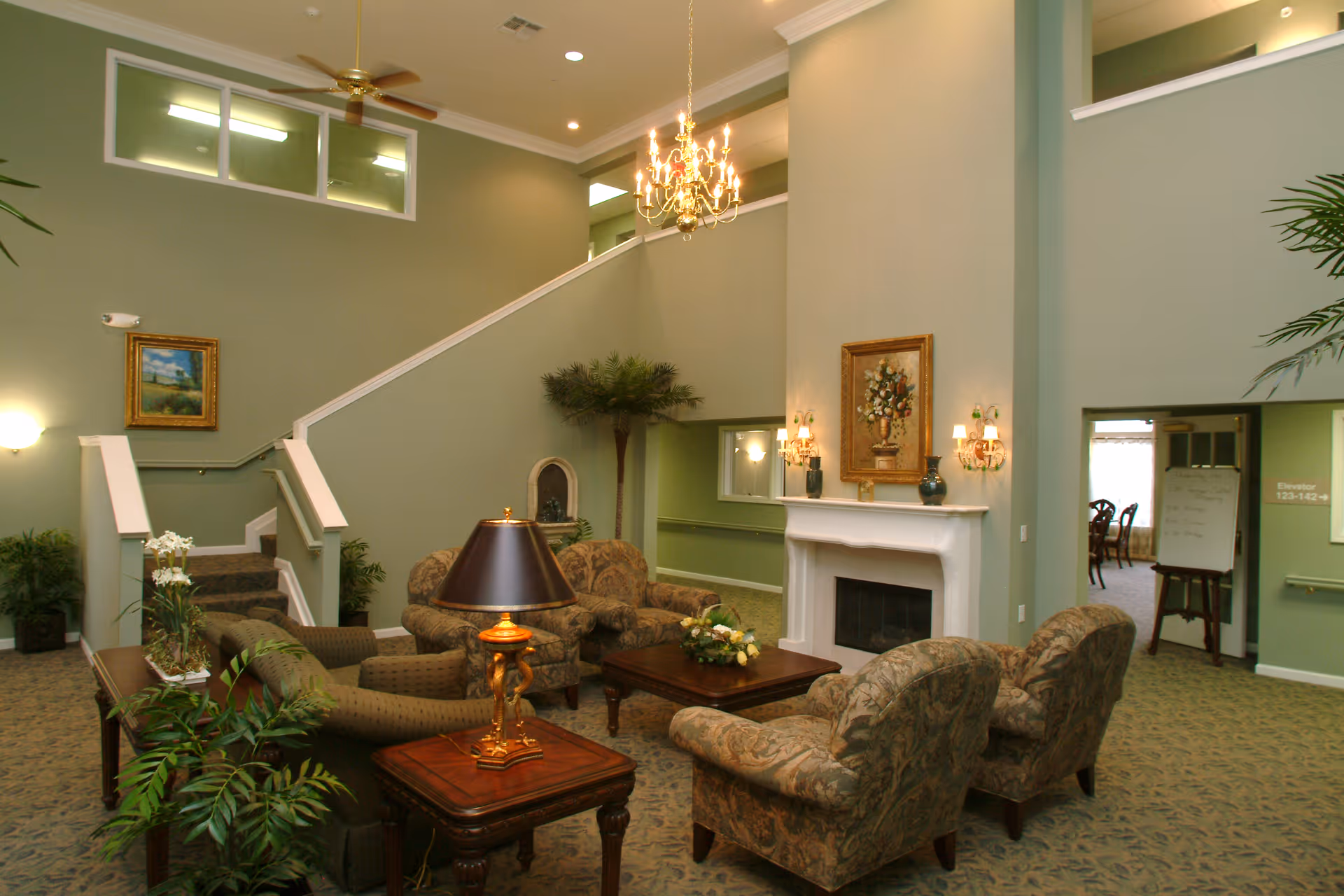 A spacious living room area in Oakhaven Manor with green walls, a white fireplace adorned with a floral painting and wall sconces, patterned armchairs and sofas arranged around a wooden coffee table with a floral centerpiece, a side table with a lamp, indoor plants, a staircase leading to an upper level, and an open doorway leading to a dining area.