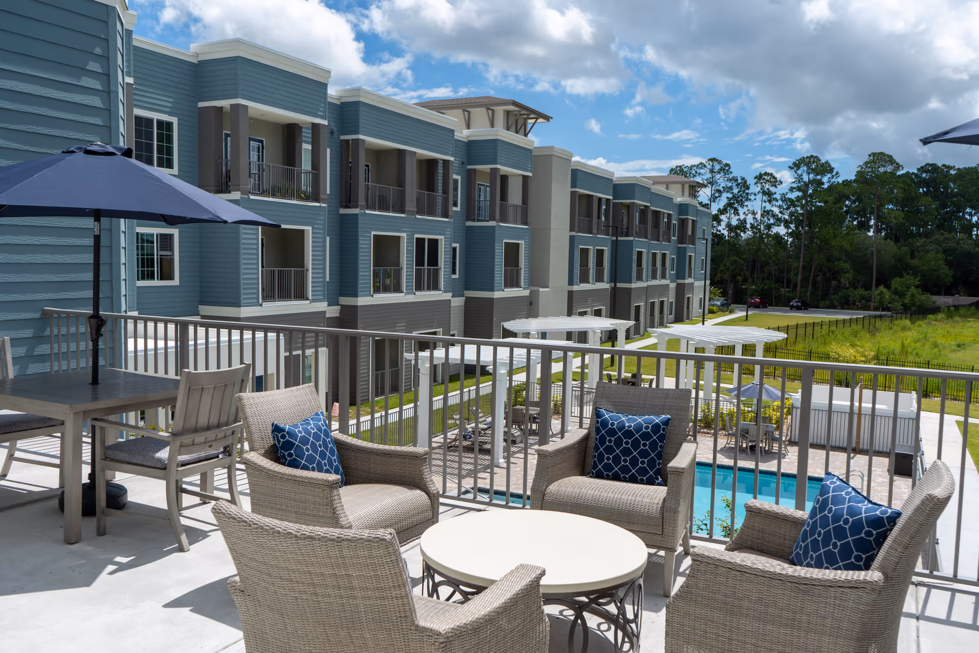 Outdoor patio area with wicker chairs and blue patterned cushions arranged around a round table, next to a table with chairs and a large blue umbrella. In the background, there is a multi-story residential building with balconies, a swimming pool, white pergolas, and a grassy area with trees under a partly cloudy sky.