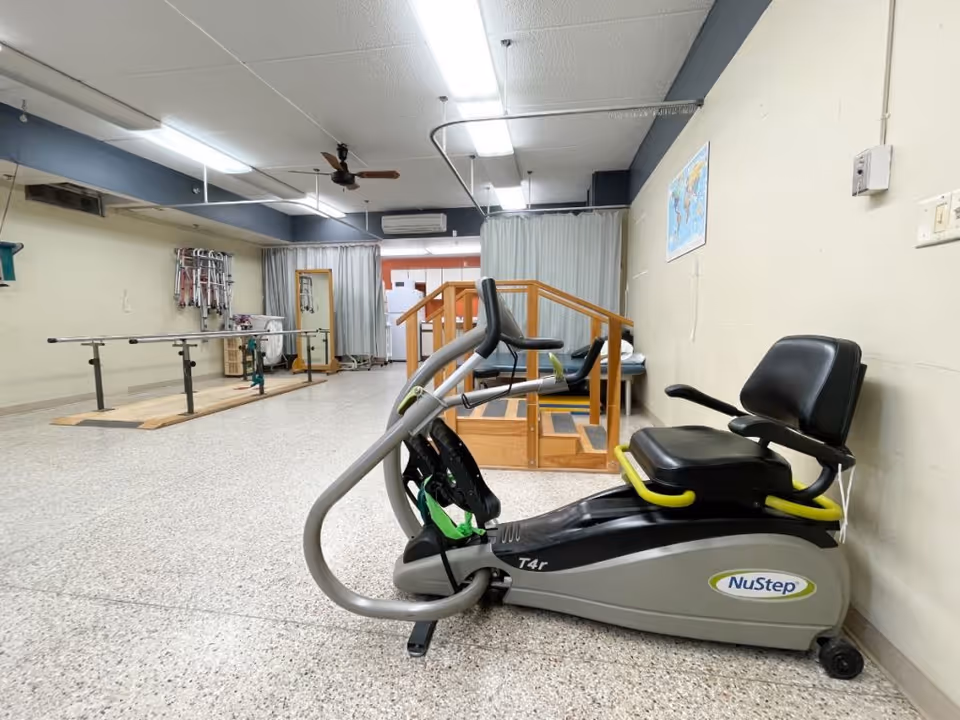 A rehabilitation or physical therapy room with a NuStep exercise machine in the foreground, parallel bars for walking practice on the left, and a wooden staircase with handrails in the background. The room has a speckled floor, fluorescent ceiling lights, and a map on the wall.