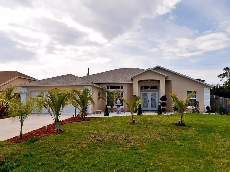 Single-story beige house with a two-car garage, a front yard with small palm trees and green grass, and a cloudy sky overhead.