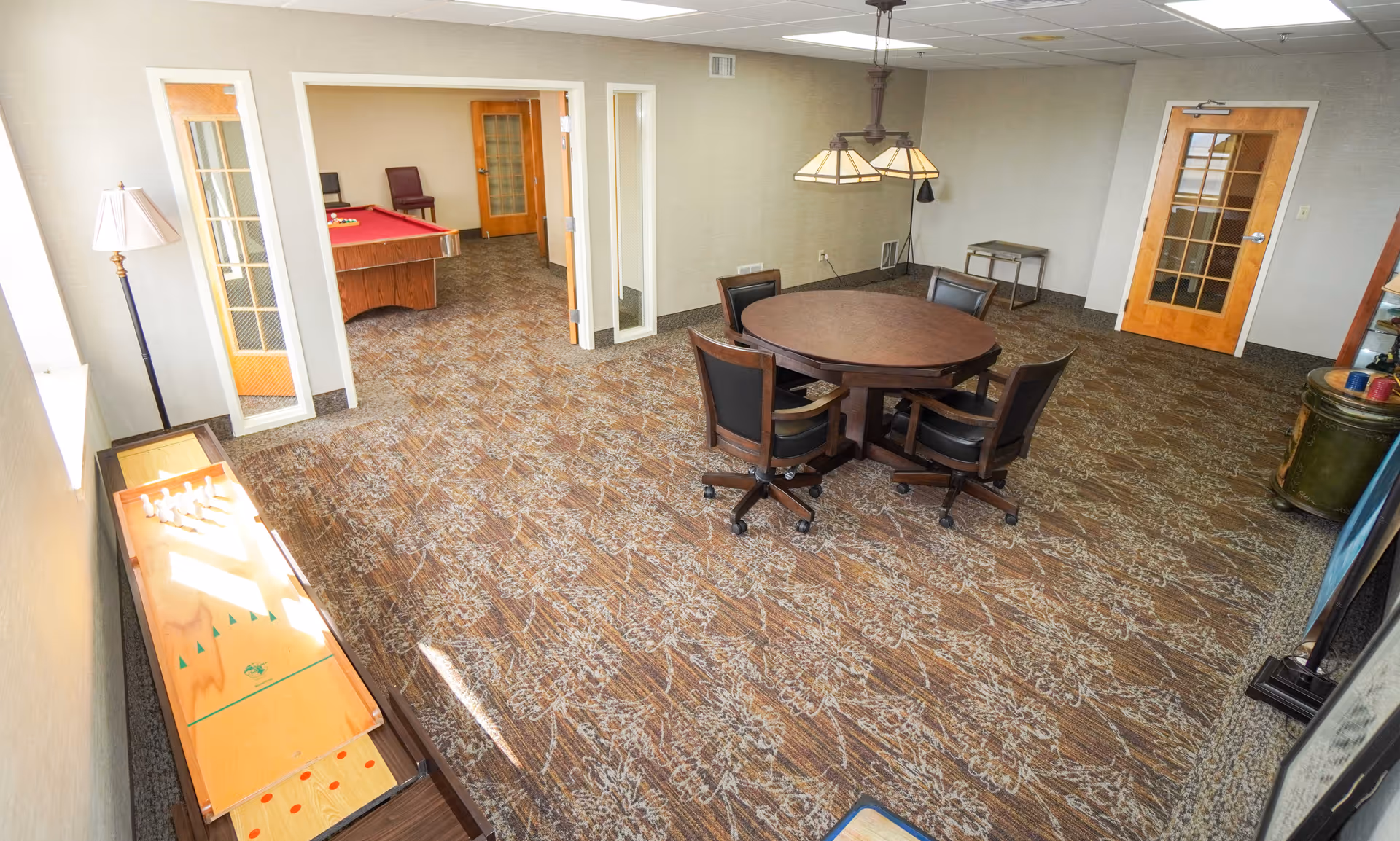Bright communal activity room with a round table and chairs, a shuffleboard to the left, and a doorway leading to a room with a pool table.