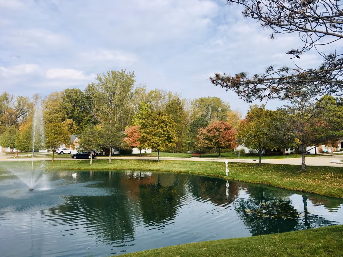 A peaceful outdoor scene at Ohio Living Swan Creek featuring a small pond with a water fountain spraying water upwards. Surrounding the pond are green grassy areas and trees with autumn-colored leaves. A few houses and parked cars are visible in the background under a partly cloudy sky.