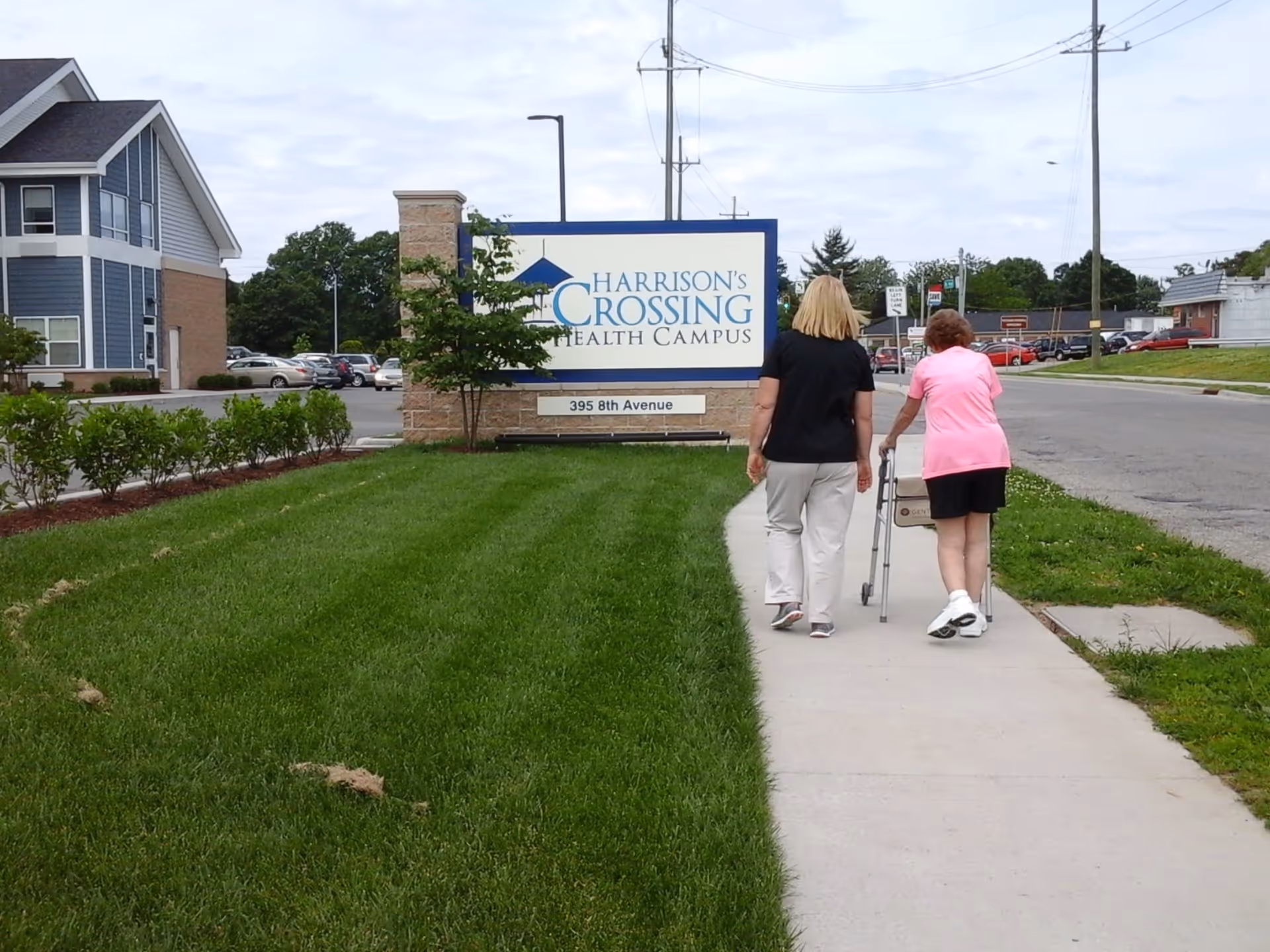 Two women walking on a sidewalk beside a green lawn towards a sign that reads 'Harrison's Crossing Health Campus, 395 8th Avenue'. One woman is using a walker and the other is walking beside her.