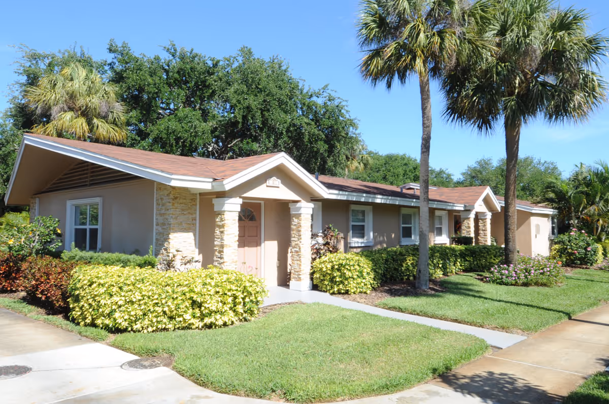 Single-story beige building with a pitched roof, palm trees and well-kept shrubs under a clear blue sky.