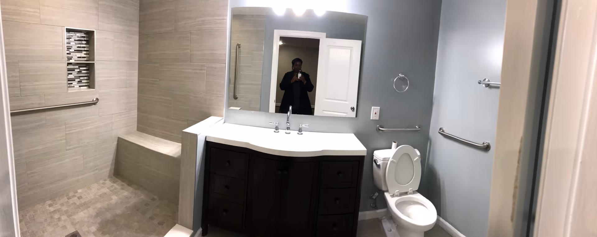 A modern bathroom with a walk-in tiled shower featuring a built-in bench and two recessed shelves with decorative tiles. There is a dark wood vanity with a white countertop and a large mirror above it. A toilet with the lid open is next to the vanity, and the walls are painted light blue with several grab bars installed for accessibility.