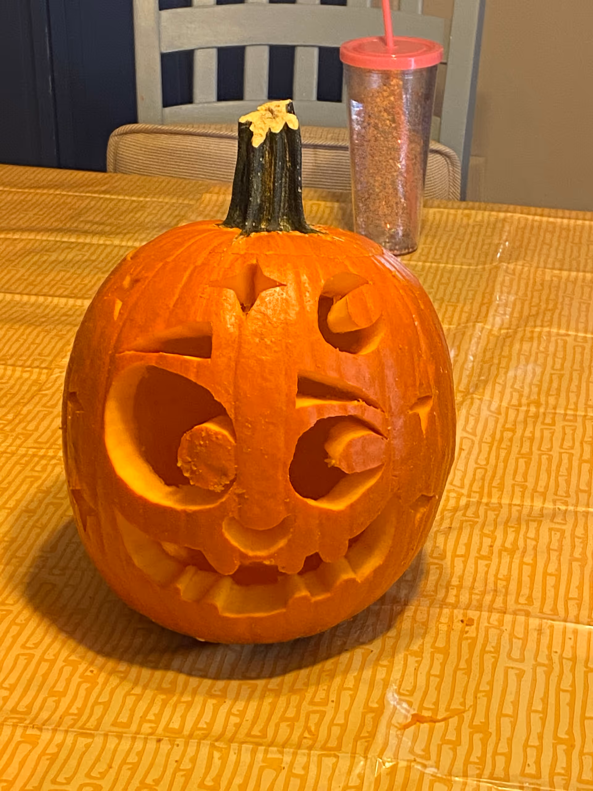 A carved pumpkin with a whimsical face featuring large eyes, a small nose, and a wide mouth with teeth, placed on a yellow patterned tablecloth. Behind the pumpkin, there is a chair and a transparent tumbler with a red lid and straw.