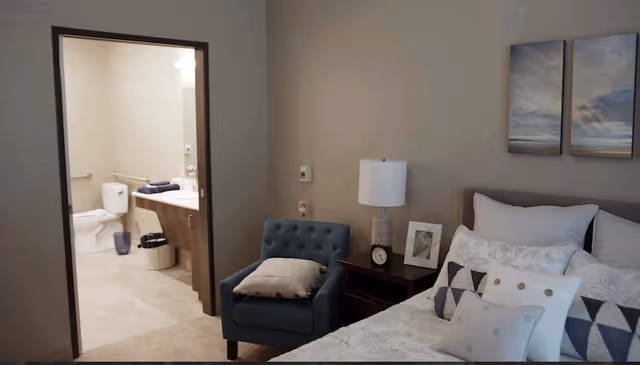 Furnished bedroom with a bed, nightstand and armchair, showing an adjacent bathroom through an open doorway.