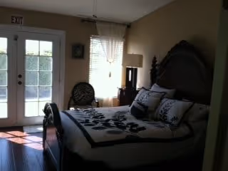 A bedroom with a large bed featuring a dark wooden headboard and white bedding with black floral patterns. There is a window with sheer curtains next to the bed, a small table with a lamp, and a chair with a patterned cushion. Double glass doors with an exit sign above them allow natural light into the room.