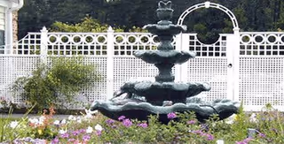 Tiered decorative fountain surrounded by flowers in a courtyard with a white lattice fence and circular trellis behind it.