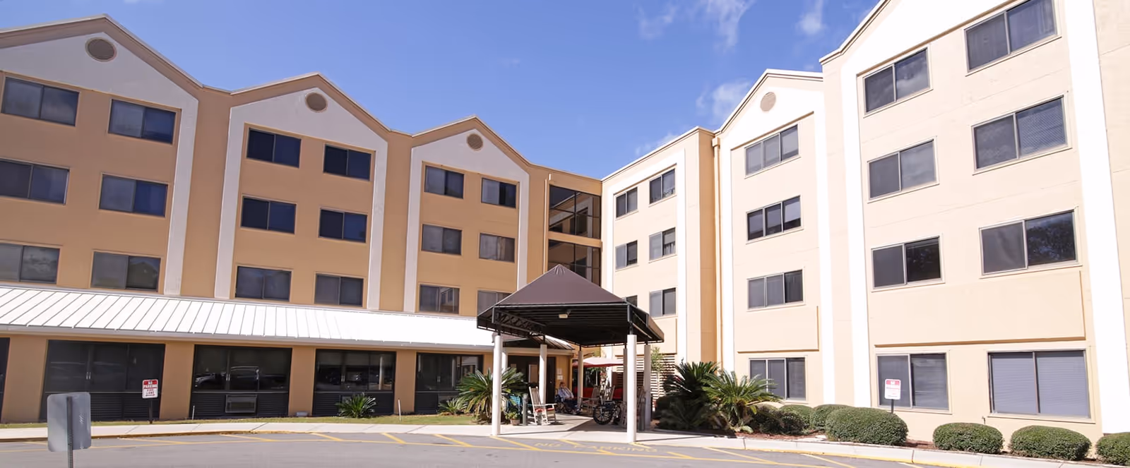 Front exterior of a multi-story beige senior living building with rows of windows and a covered entrance/porte-cochère.