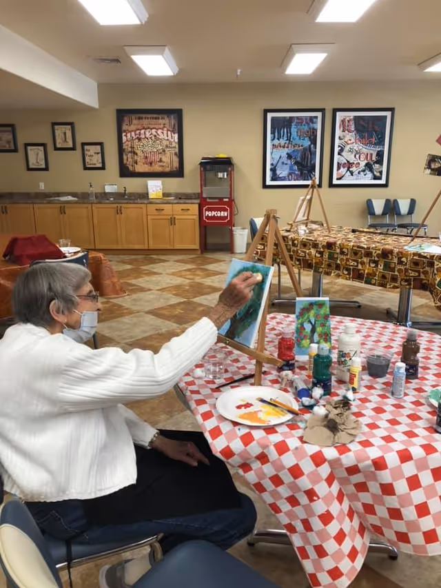 An elderly woman wearing a white sweater and face mask is seated at a table covered with a red and white checkered tablecloth, painting on a canvas set on an easel. The table has various paint bottles, brushes, and a palette with paint. The room has a tiled floor, wooden cabinets along the back wall, a popcorn machine, and framed posters on the walls.