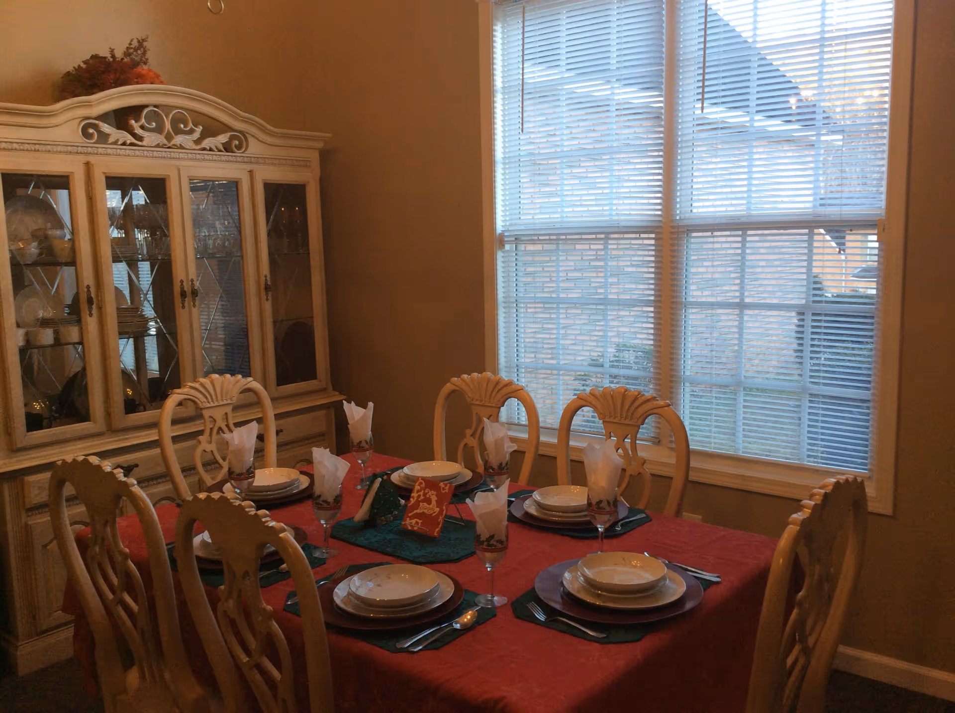 A dining room with a table set for six people. The table is covered with a red tablecloth and has plates, bowls, silverware, and glasses with folded napkins. There is a decorative centerpiece on the table. Behind the table is a large window with blinds, and to the left is a wooden china cabinet displaying dishes and glassware.