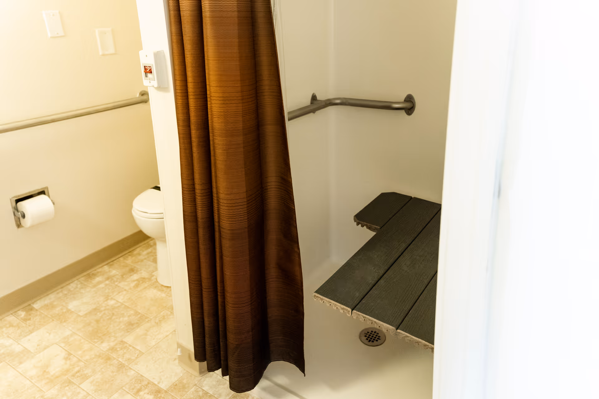 A bathroom with a toilet, a toilet paper holder, grab bars on the walls, and a shower area with a brown curtain and a fold-down shower seat.