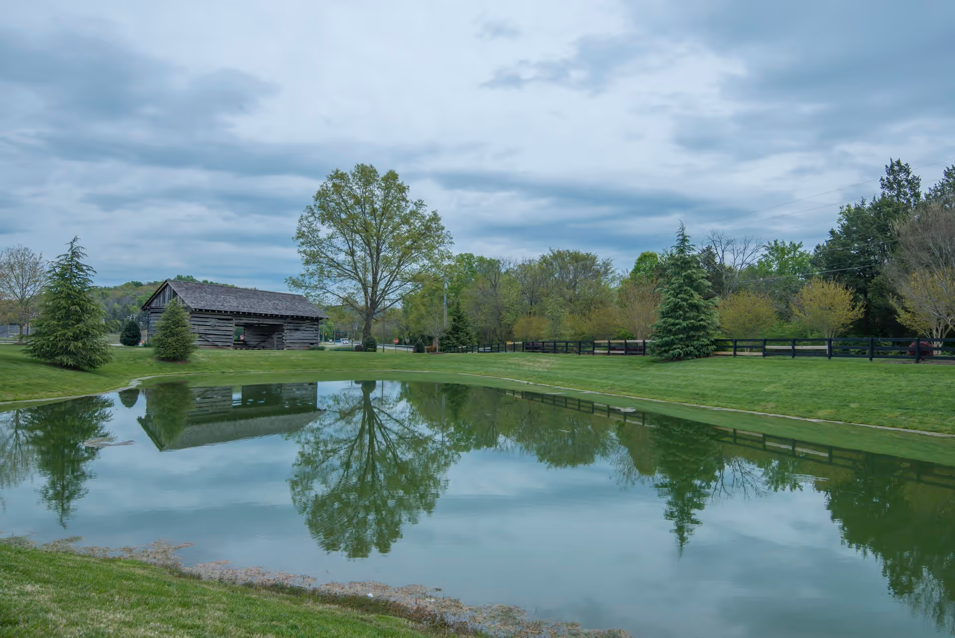 A serene outdoor scene featuring a small pond reflecting a large tree, a rustic wooden barn, and surrounding greenery including trees and grass under a cloudy sky.