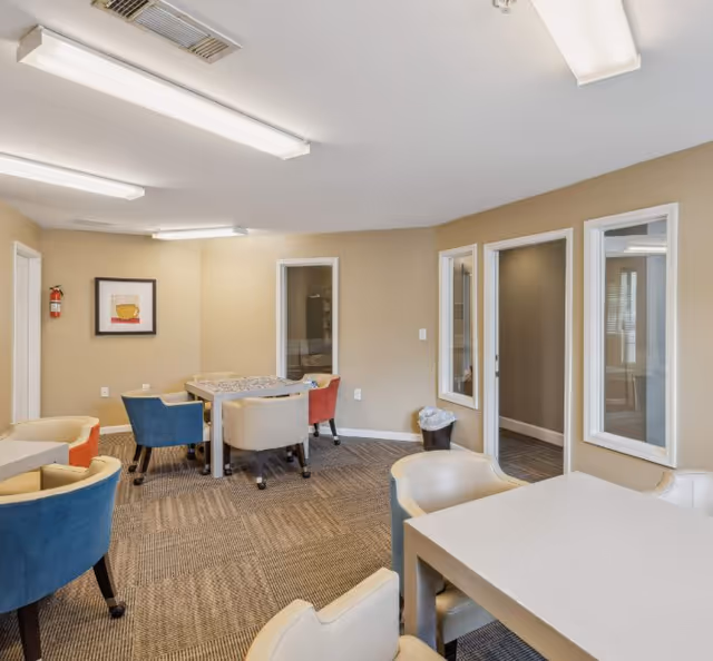 Bright communal room with tables and colorful upholstered chairs and windows into adjacent rooms.