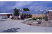 Courtyard of a single-story brick senior living facility with paved walkways, rock landscaping, benches, and a small decorative windmill under a cloudy sky.