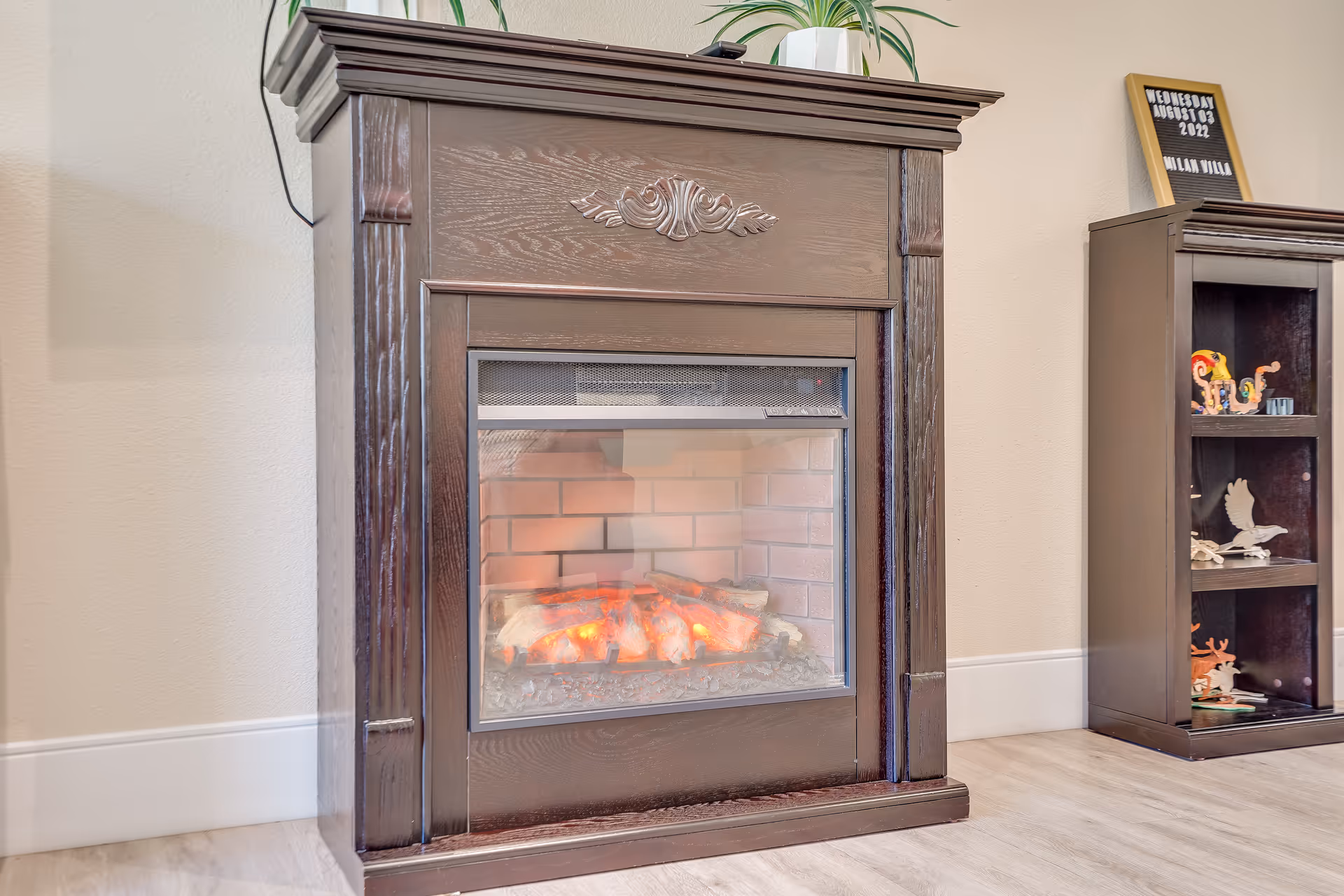 Electric fireplace with a dark wooden mantel in a living area, next to a display cabinet with decorative items.