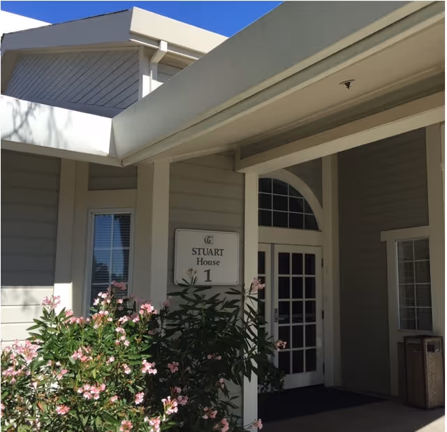Covered entrance to Stuart House 1 with glass double doors, a sign, and pink flowering shrubs.