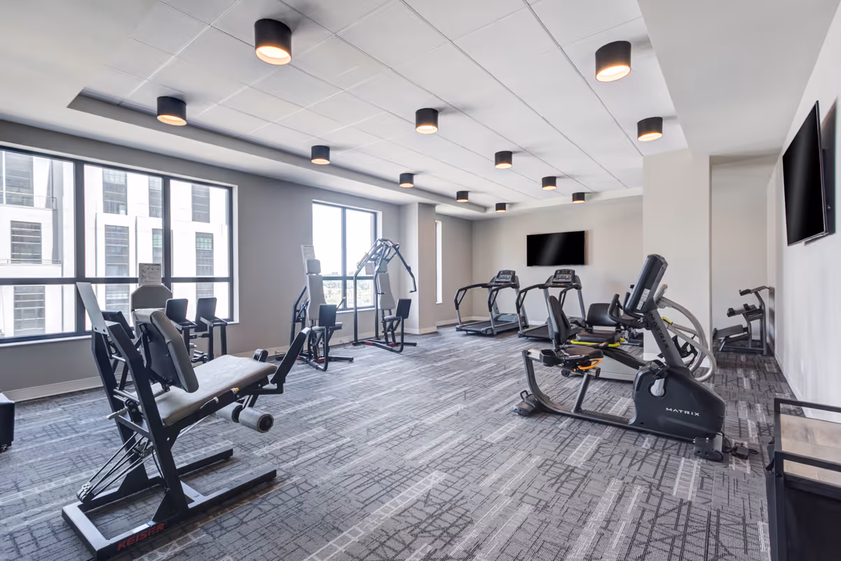A modern fitness room with various exercise equipment including stationary bikes, treadmills, and weight machines. The room has large windows allowing natural light to enter, gray walls, a patterned carpet, and multiple ceiling lights. Two flat-screen TVs are mounted on the walls.