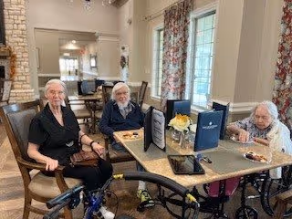Three elderly women sitting around a rectangular dining table in a well-lit room with large windows and floral curtains. The table has menus, a small flower arrangement, and plates with food. The room has a cozy and welcoming atmosphere with neutral-colored walls and wooden furniture.