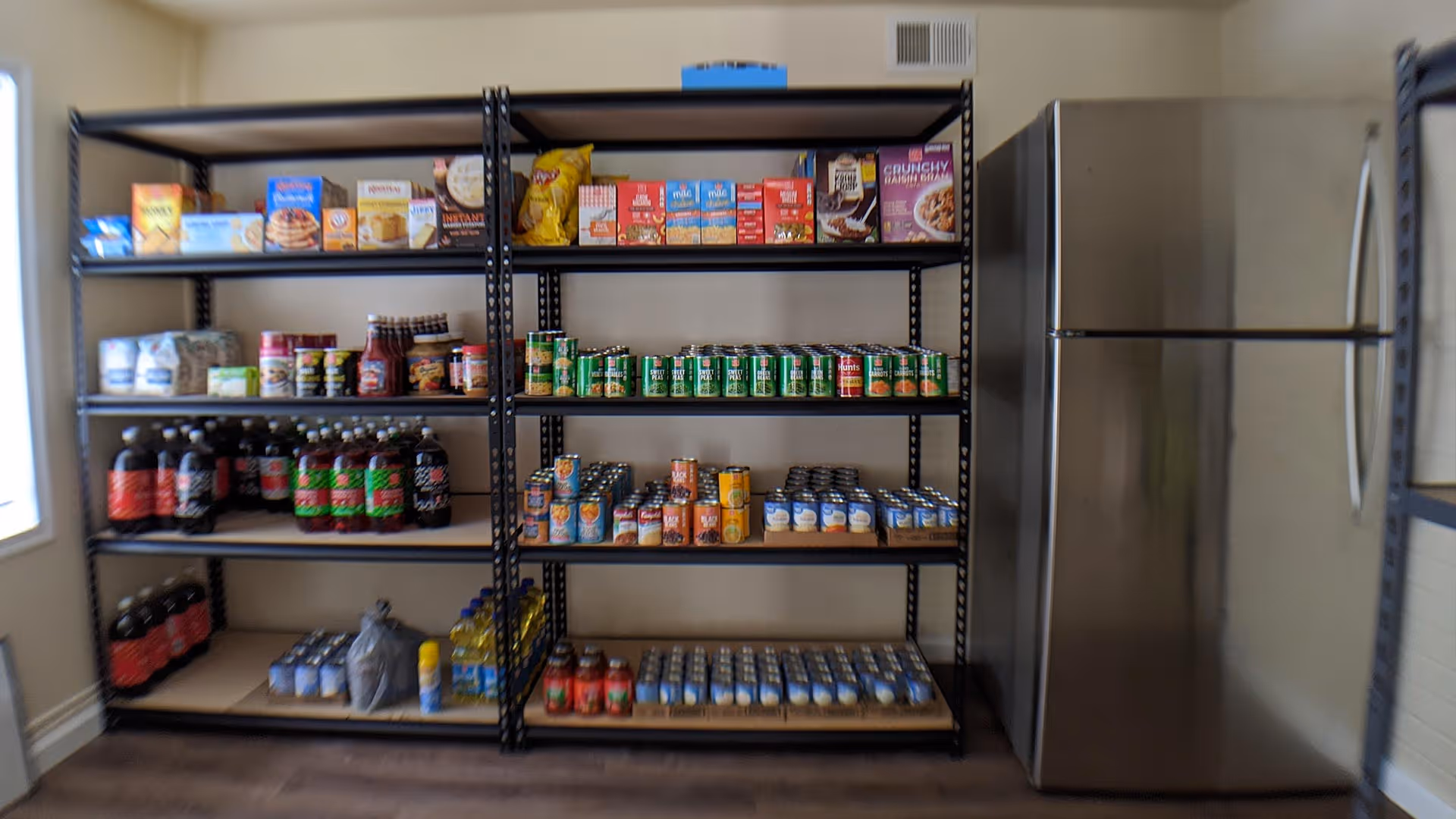 A pantry room with metal shelving units stocked with various food items including canned goods, boxed cereals, snacks, bottled drinks, and cooking oil. A stainless steel refrigerator is positioned to the right of the shelves. The room has beige walls and a window on the left side.