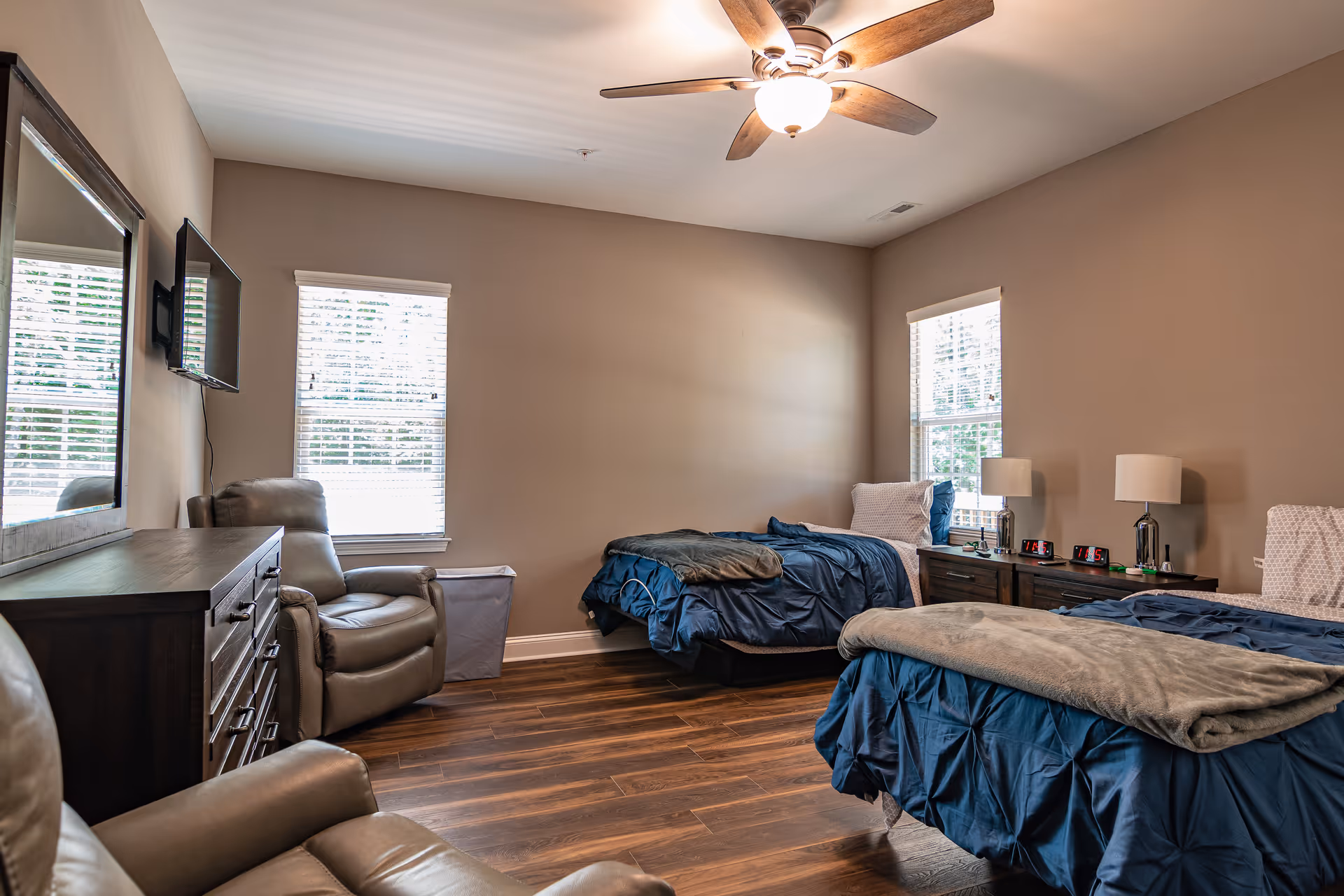 A bright two-bed bedroom with blue bedding, recliners, a dresser with wall-mounted TV, and a ceiling fan.