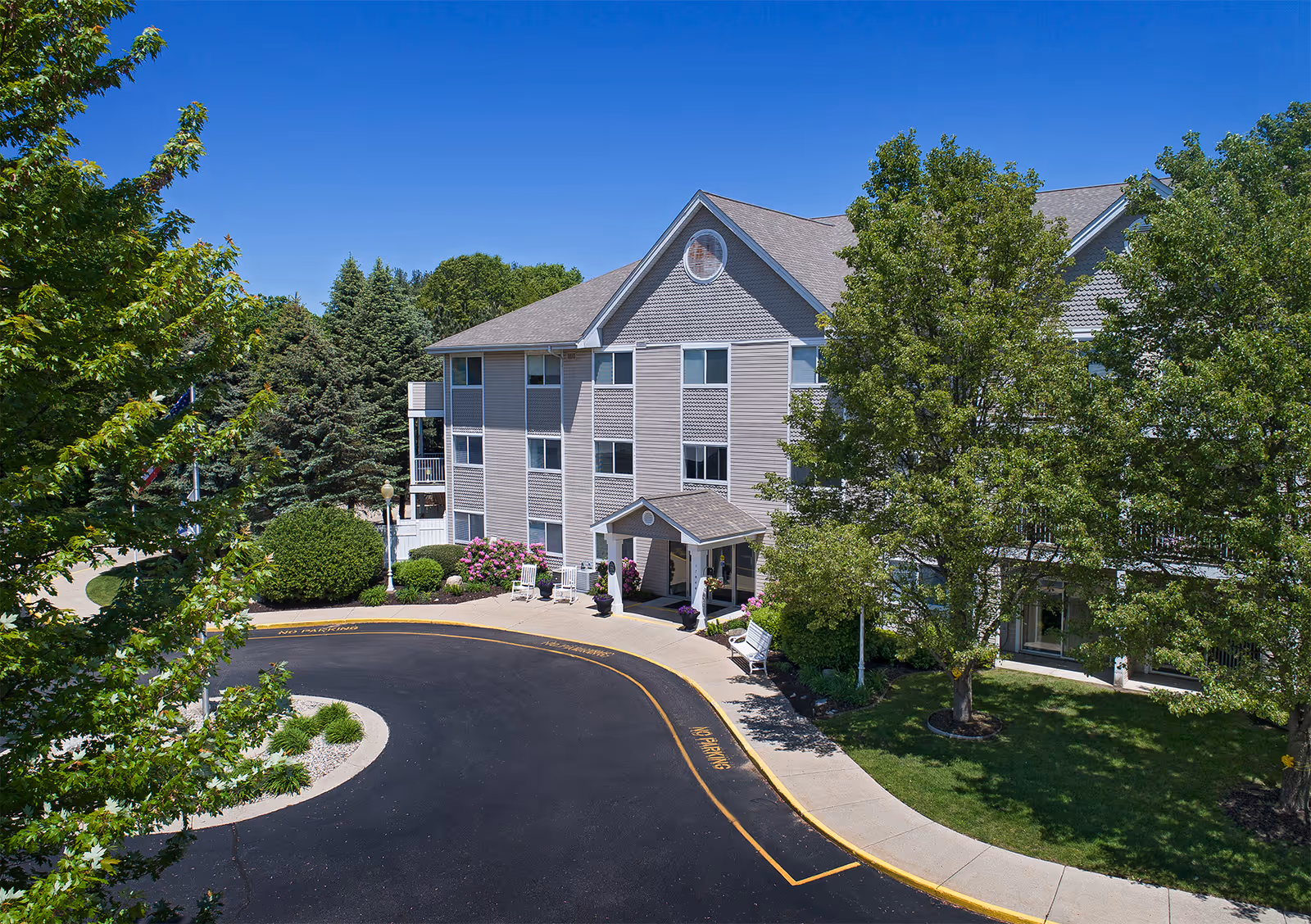 Exterior view of a three-story senior living facility building surrounded by trees and landscaping under a clear blue sky. The building has a covered entrance with benches and a circular driveway marked with no parking zones.