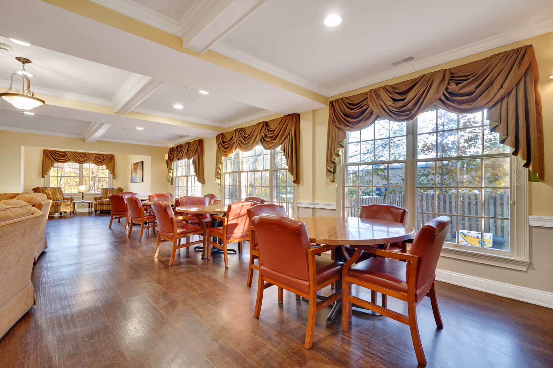 A bright and spacious dining area in a senior living facility with multiple wooden tables and red cushioned chairs arranged neatly. Large windows with brown drapes allow natural light to fill the room, and there are comfortable seating areas with sofas and armchairs in the background. The floor is wooden, and the walls are painted in a warm beige tone.