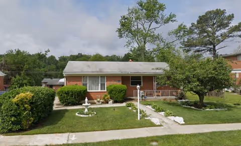 A single-story brick house with a manicured front yard, walkway, shrubs, and trees.