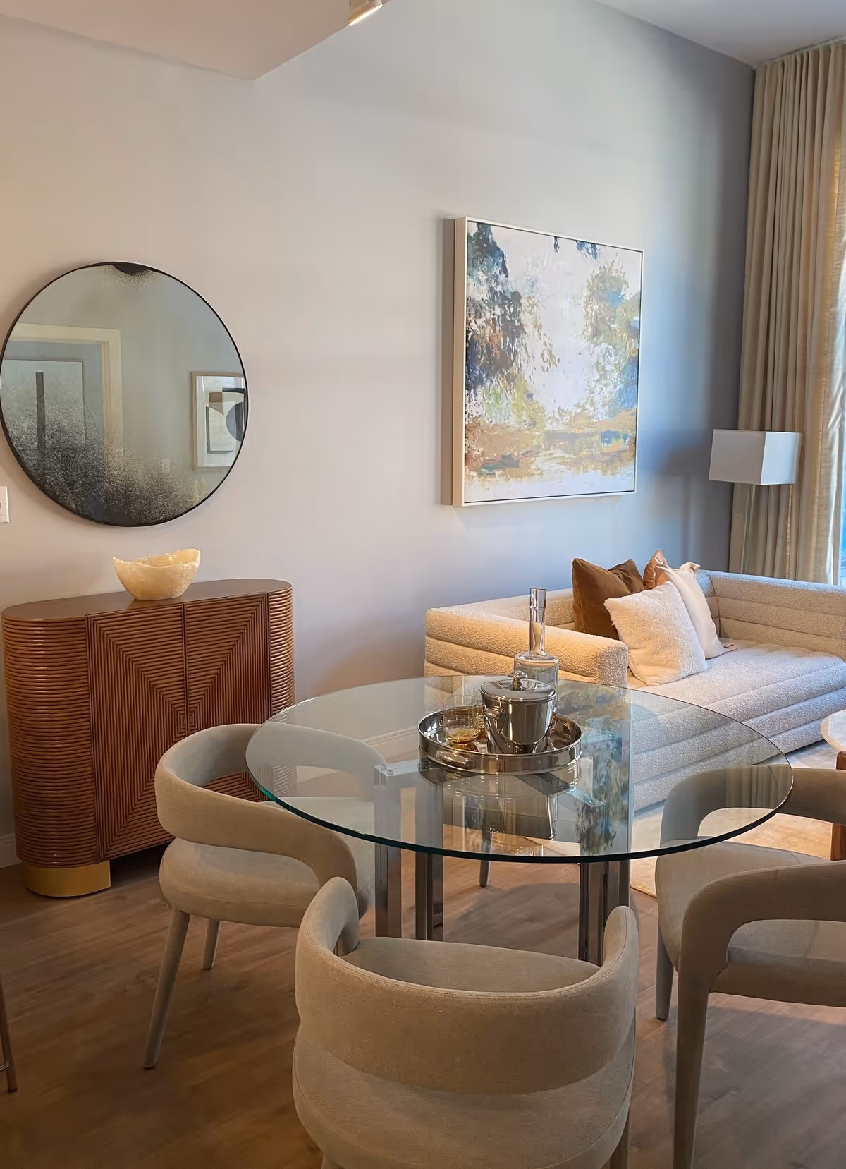 A modern living room with a round glass dining table surrounded by four beige upholstered chairs. Behind the table is a beige sectional sofa with multiple pillows, a floor lamp, and a large abstract painting on the wall. To the left, there is a wooden sideboard with a decorative bowl on top and a large round mirror hanging above it. The room has light-colored walls and wood flooring.