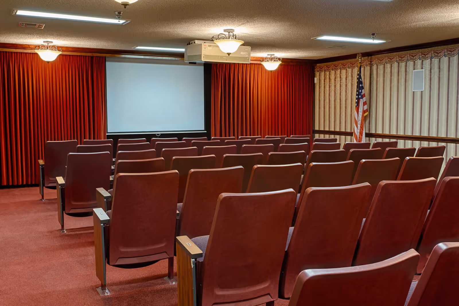Small auditorium-style screening room with rows of red upholstered seats facing a projection screen, red curtains, and an American flag.