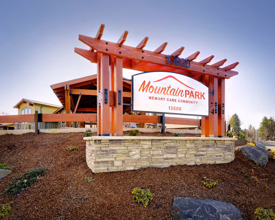 Outdoor view of a large wooden sign for Mountain Park Memory Care Community with the address 13600, set on a stone base with landscaping around it and a building in the background under a clear sky.