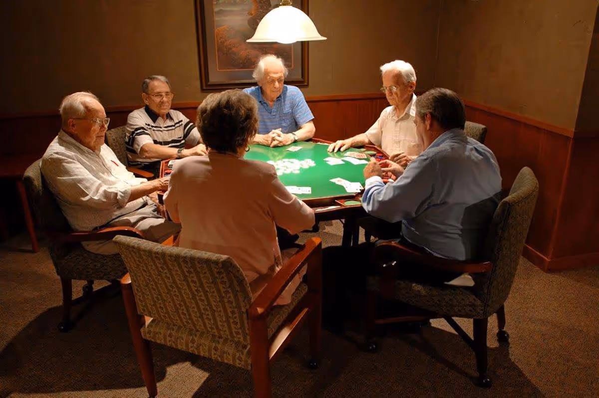 Six elderly individuals sitting around a green card table in a warmly lit room, engaged in a card game. The room has wood paneling on the walls and a framed picture hanging behind them.