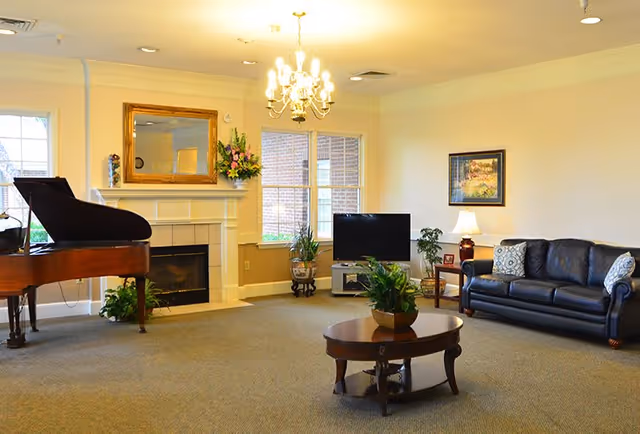 A cozy living room with a grand piano on the left, a fireplace with a large mirror and flower arrangement above it, a flat-screen TV on a stand near two windows, a black leather sofa with patterned pillows, a side table with a lamp, a framed painting on the wall, and a wooden coffee table with a plant in the center.