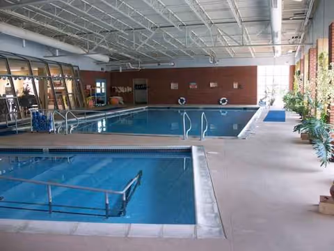 Indoor nursing home pool area with two pools, rails, lifebuoys, and potted plants.