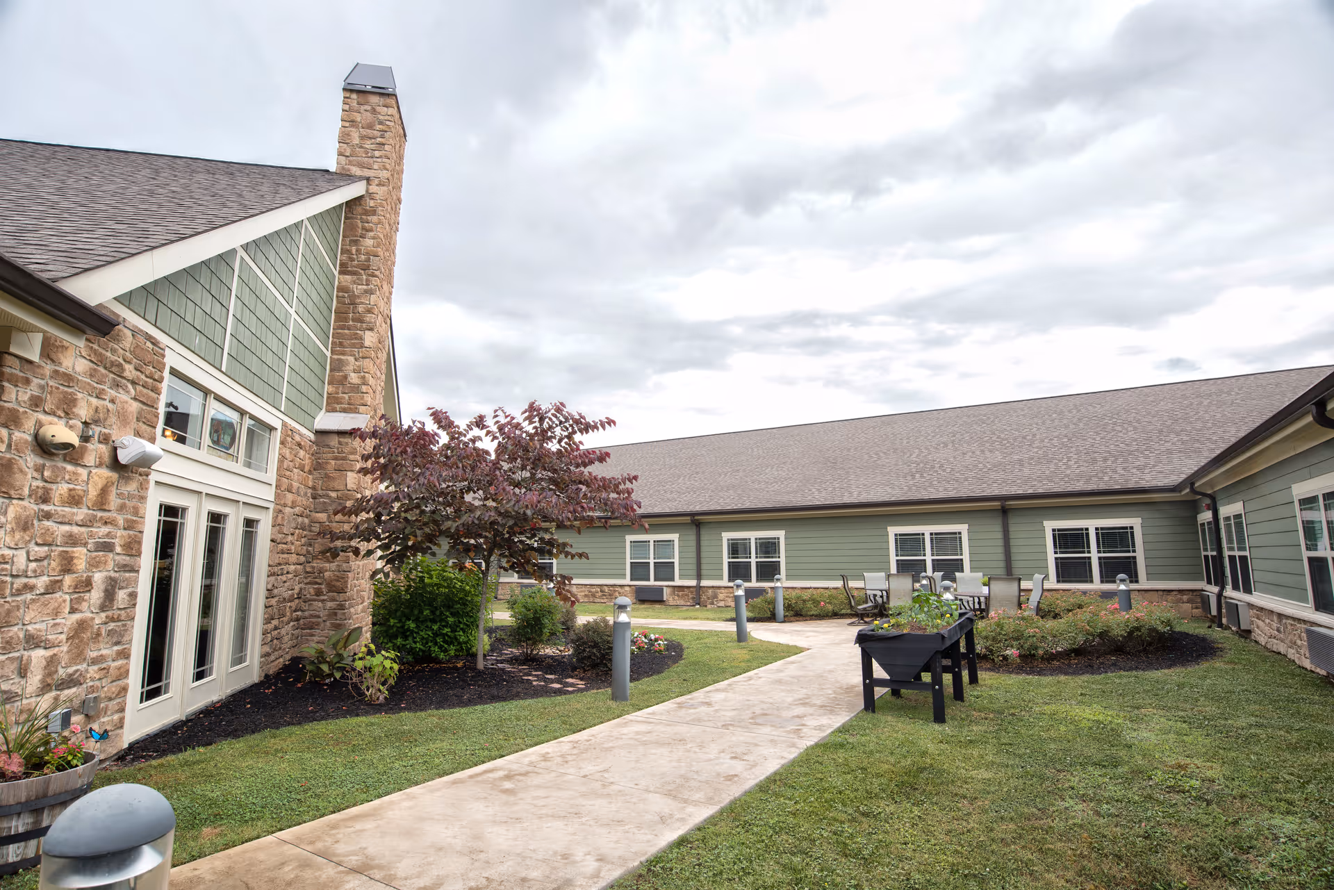 Outdoor courtyard area of a senior living facility with a paved walkway, green grass, small trees, and shrubs. The building has stone and green siding with multiple windows and a chimney. There are outdoor tables and chairs along the walkway under a cloudy sky.