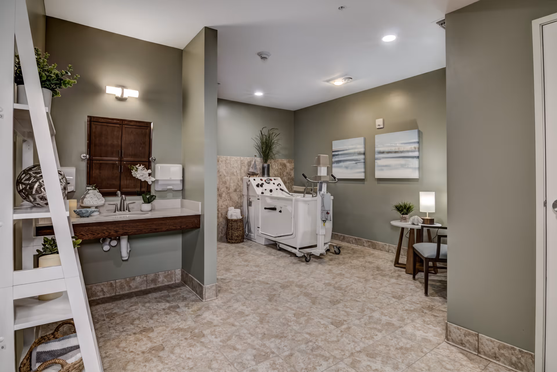 A clean and well-lit room with beige tiled flooring and muted green walls. On the left side, there is a white shelving unit with decorative items and plants, next to a wooden vanity with a sink, faucet, and a mirror above it. In the center and towards the back of the room, there is a white assisted bathing tub with controls and a chair attached. On the right side, there is a small round table with a lamp, plant, and chair. Two abstract paintings hang on the wall above the table.