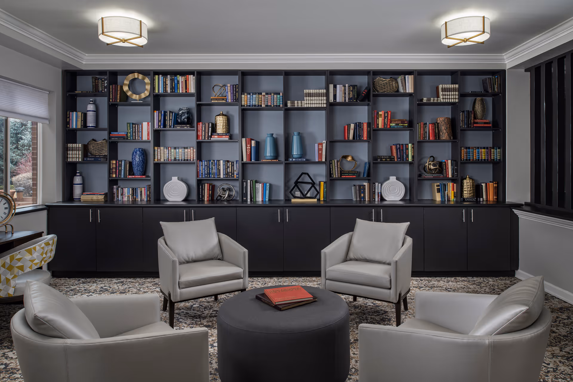A cozy reading or lounge area featuring four light gray armchairs arranged around a round dark gray ottoman with books on top. Behind the chairs is a large black bookshelf filled with books and decorative items. The room has a patterned carpet, two ceiling lights, and a window with a view of trees outside.