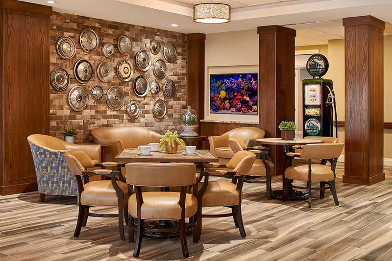 Seating area with round tables and leather chairs in a senior living common room featuring a decorative hubcap wall, an aquarium, and wooden columns.